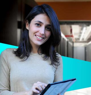 Female IT worker holding laptop in server room and smiling at the camera.