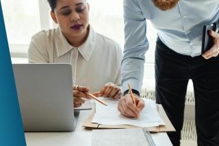 Two professional recruitment consultants collaborating over a detailed market intelligence report and salary benchmarking data to provide a client with accurate pay-scale insights for cyber security roles.