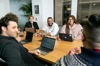 A team with laptops around a meeting table listening to a colleague