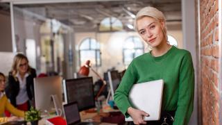 Professional woman in workplace holding laptop leaning against a brick wall.